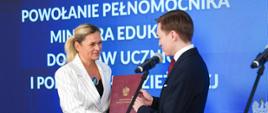 Ministra Edukacji Barbara Nowacka wręcza akt powołania w czerwonej teczce Mikołajowi Wolaninowi na tle ścianki z napisem informacyjnym.