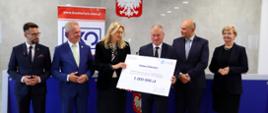 Pod szarą ścianą przed niebieskim stołem stoi sześć osób, kobieta i mężczyzna pośrodku trzymają wielki symboliczny czek z napisem Gmina Zakrzew - 1 000 000 zł.