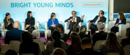 Na tle jasnoniebieskiej ściany z napisem Bright Young Minds w białych fotelach siedzi kilka osób, mężczyzna w szarym garniturze i okularach mówi do mikrofonu, przed nimi widać sale na której na krzesłach siedzi wiele osób i słucha.