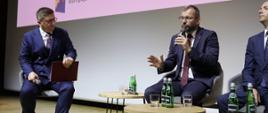 Minister Grzegorz Puda siedzi przy stole i przemawia do mikrofonu