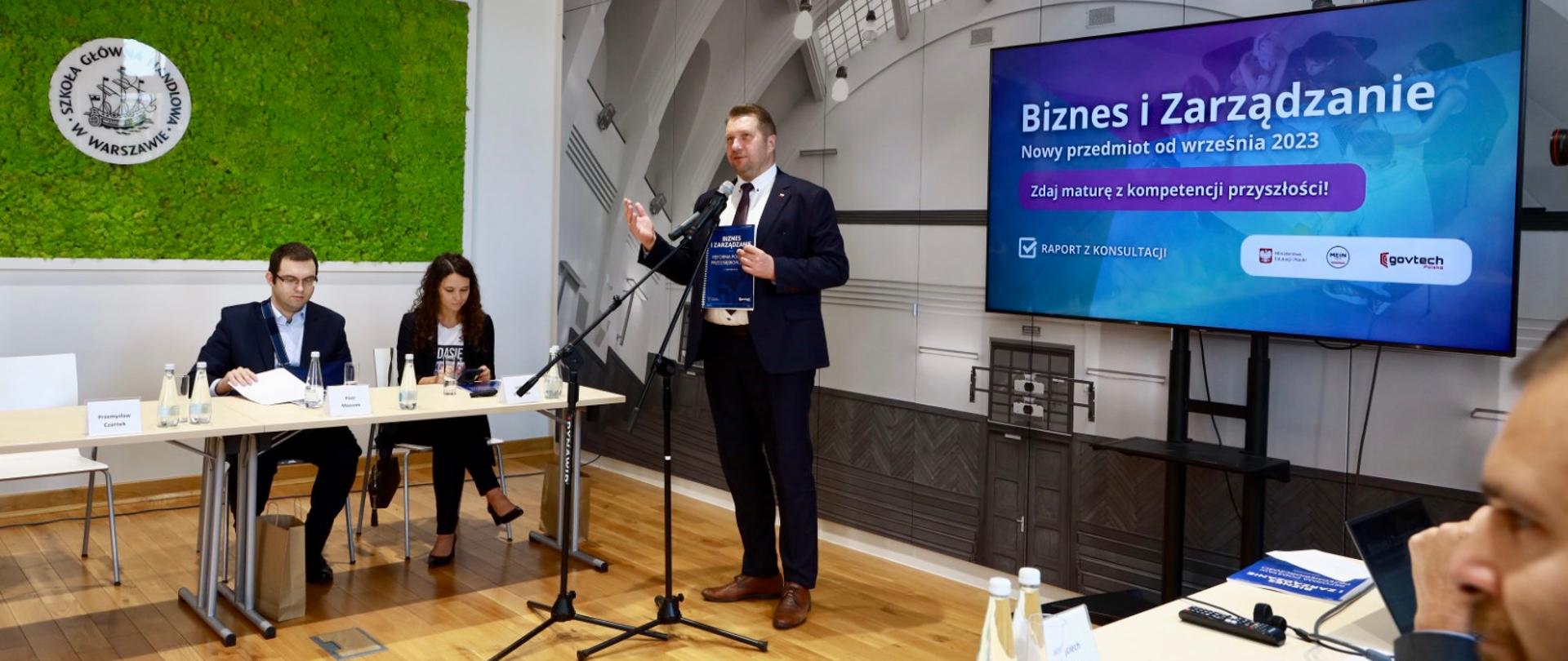 Mężczyzna w czarnym garniturze stoi ii mówi do mikrofonu na stojaku, za nim duży monitor z napisem Biznes i zarządzanie na niebieskim tle.