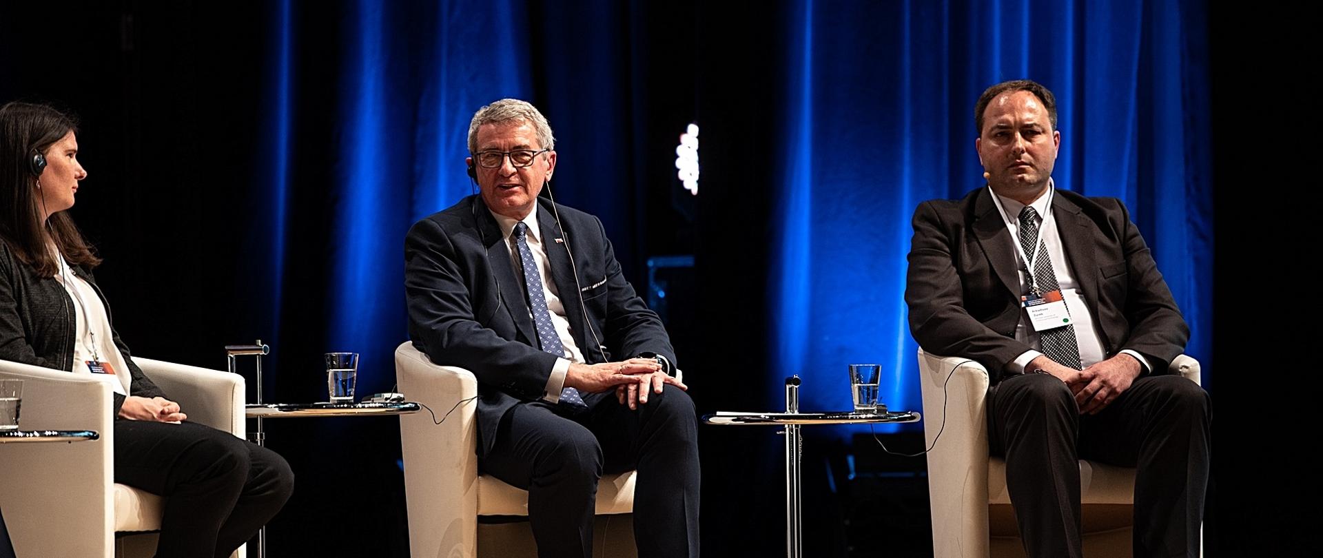 Na białych fotelach siedzą kobieta w czarnej marynarce, wiceminister Murdzek i mężczyzna w czarnym garniturze, za nim zasłonięta granatową kotarą ściana.