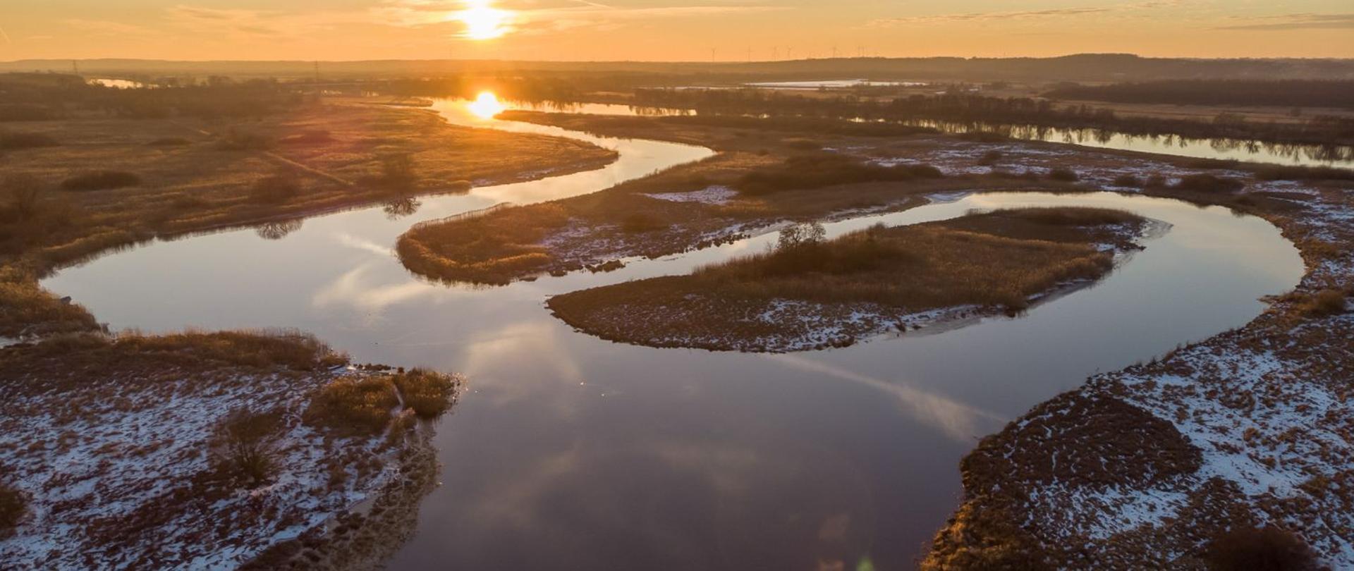 Na zdjęciu widoczna jest rzeka Odra. Na brzegach rzeki leży delikatny śnieg. W tle widać zachodzące słońce.