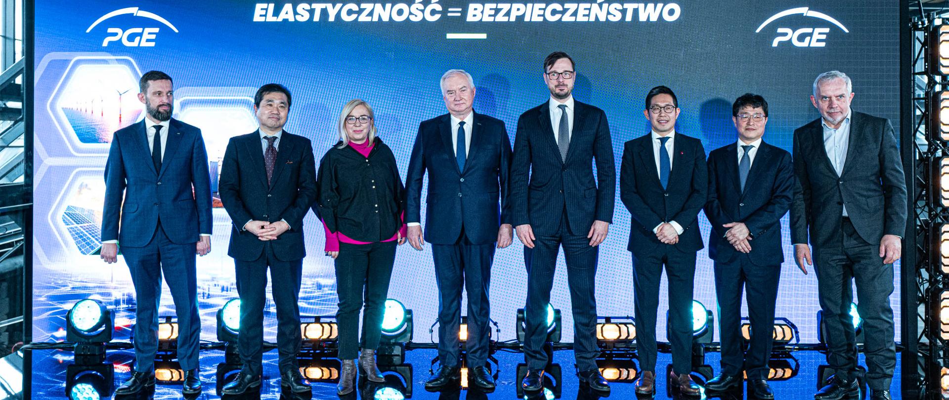 Ministra Paulina Hennig-Kloska stoi w rzędzie z przedstawicielami Polskiej Grupy Energetycznej i firm biorących udział w spotkaniu organizowanym przez PGE. Osoby pozują, stojąc na tle niebieskiego ekranu z napisem cytuję: elastyczność = bezpieczeństwo.