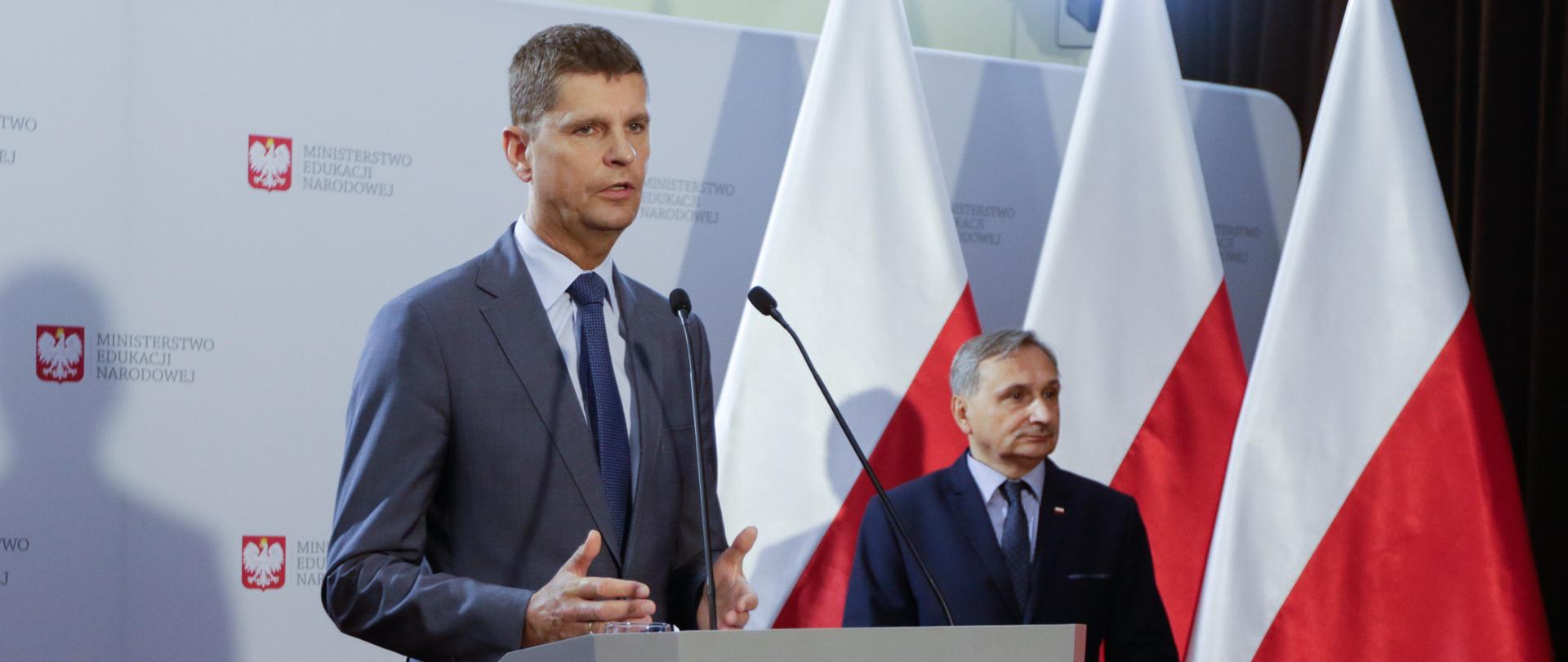 Minister Edukacji Narodowej Dariusz Piontkowski i wiceminister Maciej Kopeć podczas konferencji. W tle biała ścianka z logo ministerstwa oraz flagi Polski.