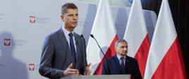Minister Edukacji Narodowej Dariusz Piontkowski i wiceminister Maciej Kopeć podczas konferencji. W tle biała ścianka z logo ministerstwa oraz flagi Polski.