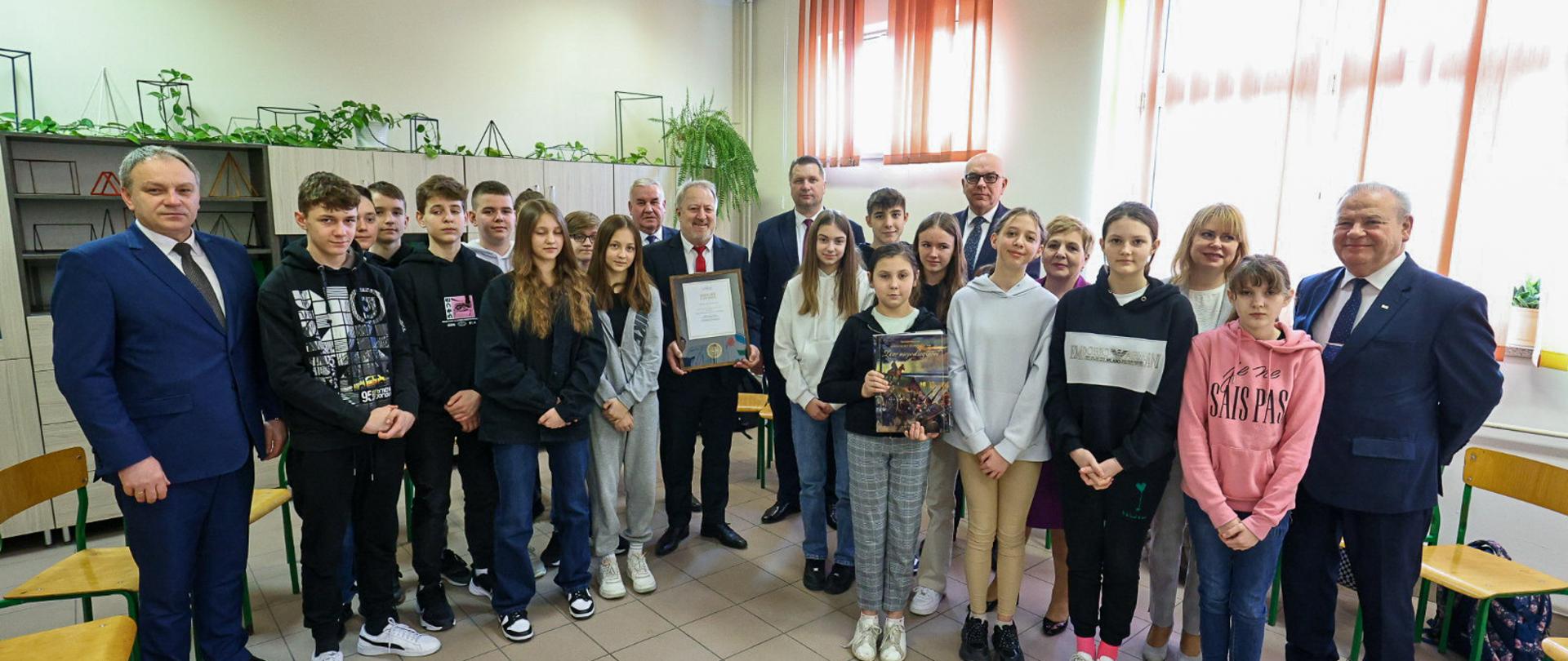 W szkolnej klasie na podłodze z jasnoszarych kafli stoi grupa młodzieży i kilkoro dorosłych, za nimi półki na ścianie, z prawej zasłonięte czerwonymi roletami okna.