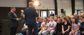 Widok z przodu na wypełnioną kolorowo ubranymi dziećmi salę, przed nimi stoi mężczyzna w garniturze i mówi do trzymanego w ręku mikrofonu, obok niego stoi wiceminister Rzymkowski.