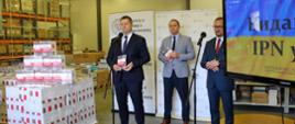 Minister Czarnek i kilka osób stoją w hali magazynowej, przed nimi mikrofony na statywach.
