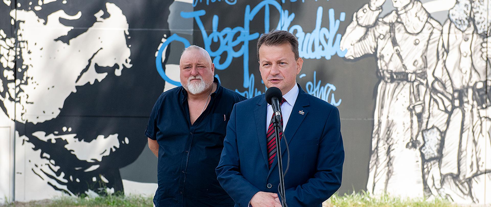 Minister Mariusz Błaszczak na tle muralu patriotycznego w Legionowie. Na murze okalającym budynek widoczny Józef Piłsudski. Obok szefa MON autor muralu.