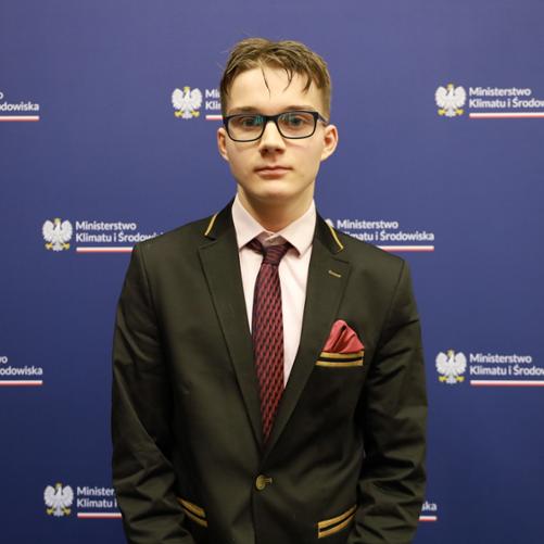 Na zdjęciu znajduje się młody mężczyzna w okularach. Ubrany jest w białą koszulę oraz czarną marynarkę. Na szyi ma zawiązany krawat, w kieszeni marynarki jest czerwona poszetka. Kieszenie zdobione są złotymi paskami.