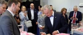 W sali z białymi ścianami stoi kilkanaście osób, na środku szpitalne łóżko na którym na żółto-różowej pościeli leży manekin dziecka, pochyla się nad nim mężczyzna w czarnym garniturze.