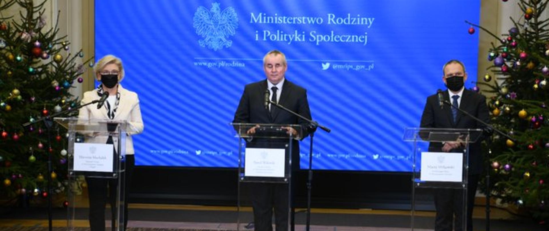 Na tle logo Ministerstwa Rodziny i Polityki Społecznej przy mównicach stoją trzy osoby. W centralnej części wiceminister Wdówik. Przy jego nogach leży czarny lablador.