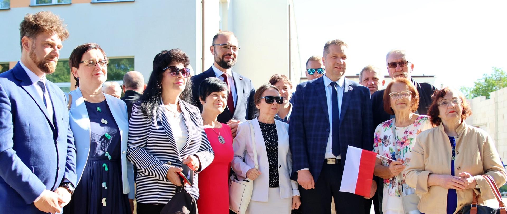 W ostrym słońcu przed białym budynkiem stoi kilkanaście osób.