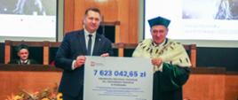 Minister Czarnek stoi obok mężczyzny w zielonym ceremonialnym stroju i zielonej czapce, trzymają wielki symboliczny czek z napisem 7 623 042,56 zł.