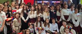 Zdjęcie zbiorowe, w dużej sali stoi grupa elegancko ubranych dzieci, za nimi dorośli, za nimi na ścianie napis Opowiedz mi, moja ojczyzno, jak matka dzieciom ciekawym...