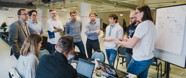 Uczestnicy hackathonu stoją i dyskutują. W tle tablica z odręcznymi notatkami.
