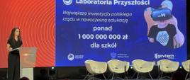 Przed zaciemnioną salą, w której w rzędach siedzą ludzie, stoi ciemno ubrana kobieta i mówi do mikrofonu, za nią kolorowa ściana z napisem Laboratoria przyszłości.