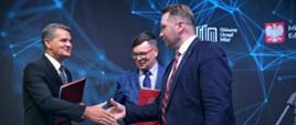 Minister podaje rękę mężczyźnie w garniturze, trzymającemu czerwoną teczkę, za nimi stoi trzeci - też z czerwoną teczką.