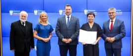 Pod ścianą stoi grupa pięciu osób, pośrodku minister Czarnek, stojąca obok niego kobieta w czarnym ubraniu trzyma otwartą teczkę z dokumentami.