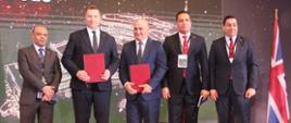 Pięciu mężczyzn w garniturach stoi na błyszczącej podłodze, dwóch trzyma czerwone teczki.