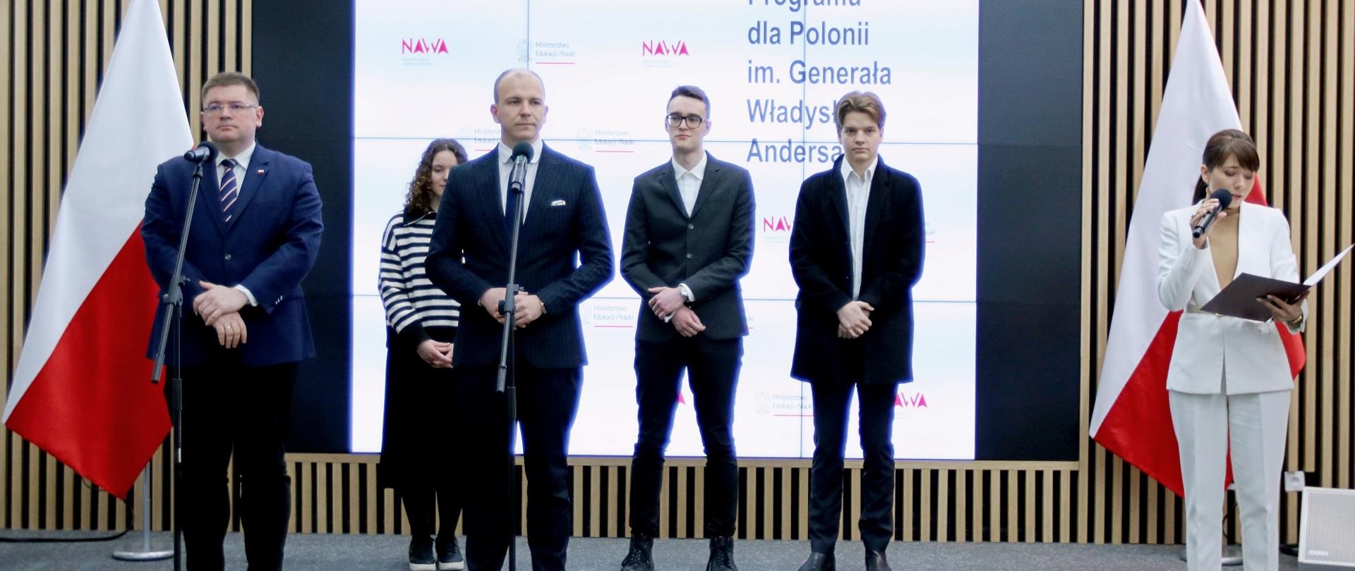 Na tle wielkiego ekranu z napisami NAWA i z polskimi flagami po bokach stoi grupa ludzi, z frontu dwóch mężczyzn w garniturach przy mikrofonach na stojakach, z boku ubrana na biało kobieta mówi do trzymanego w ręku bezprzewodowego mikrofonu.