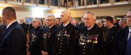 Widok na dużą salę, przy ustawionych w rzędy krzesłach stoją mężczyźni w czarnych galowych mundurach, na których mają różne medale na kolorowych wstążkach.