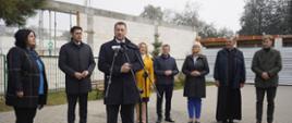Na wyłożonym szarą kostką placyku stoi minister Czarnek i mówi do mikrofonu na stojaku, za nim stoi kilka osób, w tle rozpoczęta budowa.