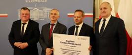 Pod szarą ścianką z napisem Śląski Urząd Wojewódzki w Katowicach stoi czterech mężczyzn w garniturach, jeden pośrodku grupy trzyma białą tabliczkę z napisem Branżowe Centrum Umiejętności.