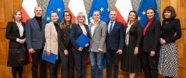 Grupa kobiet i mężczyzn pozuje do zdjęcia. W tle trzy flagi biało-czerwone i trzy flagi niebieskie z żółtymi gwiazdkami.