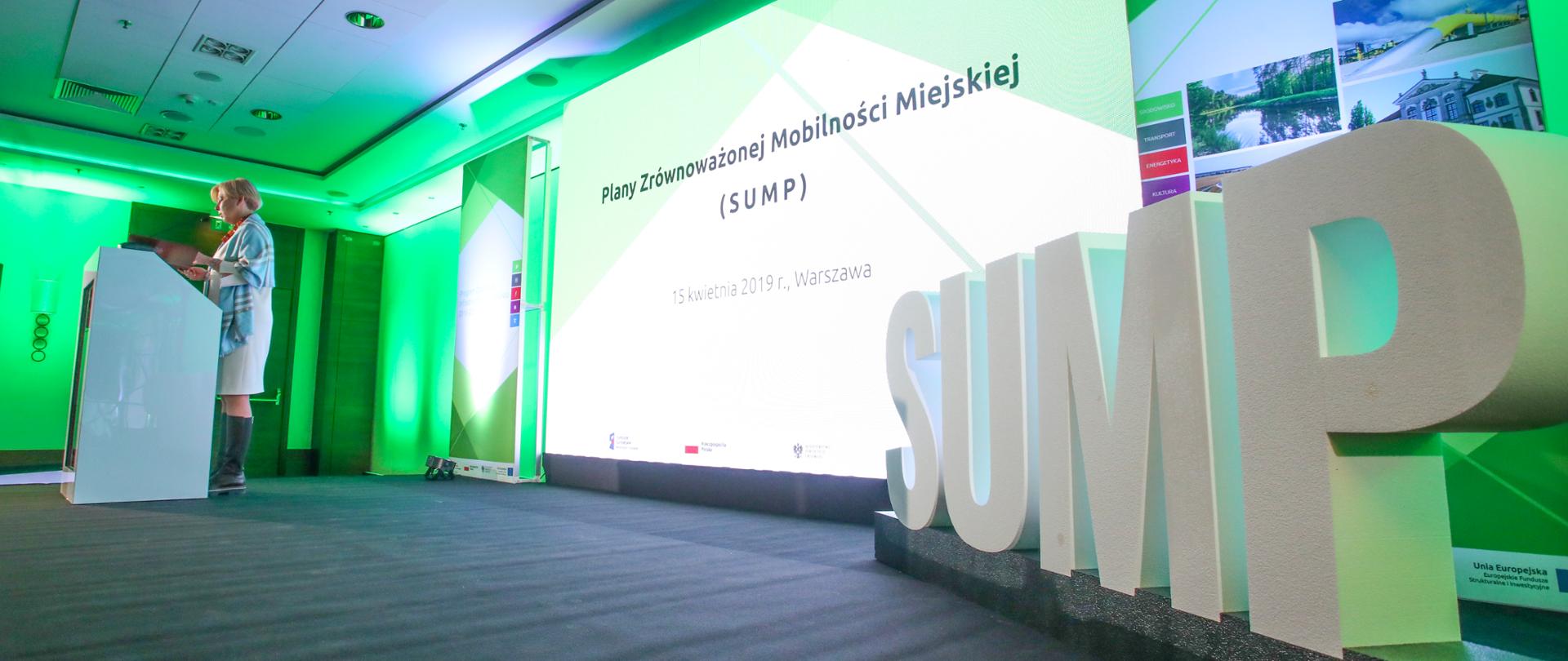 Minister Zielińska stoi przy mównicy. Za nią znajduję się tablica na której wyświetlany jest napis "Plany Zrównoważonej Mobilności Miejskiej (SUMP). 15 kwietnia 2019 r., Warszawa. Z boku stoi napis "SUMP". 
