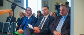 Widok z boku, w pierwszym rzędzie krzeseł siedzi minister Czarnek i kilku mężczyzn w garniturach, za nimi inni ludzie, dalej w jasnej ścianie dwa okna.