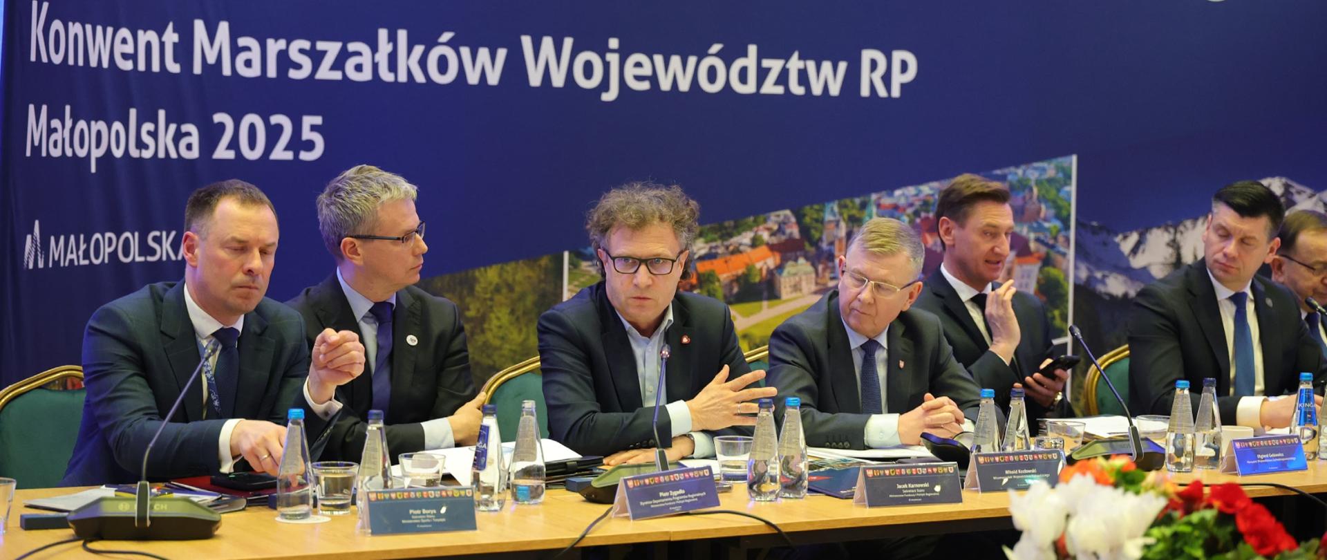 Siedem osób siedzi obok siebie przy drewnianym stole. Trzeci od lewej siedzi wiceminister Jacek Karnowski i mówi do mikrofonu. 