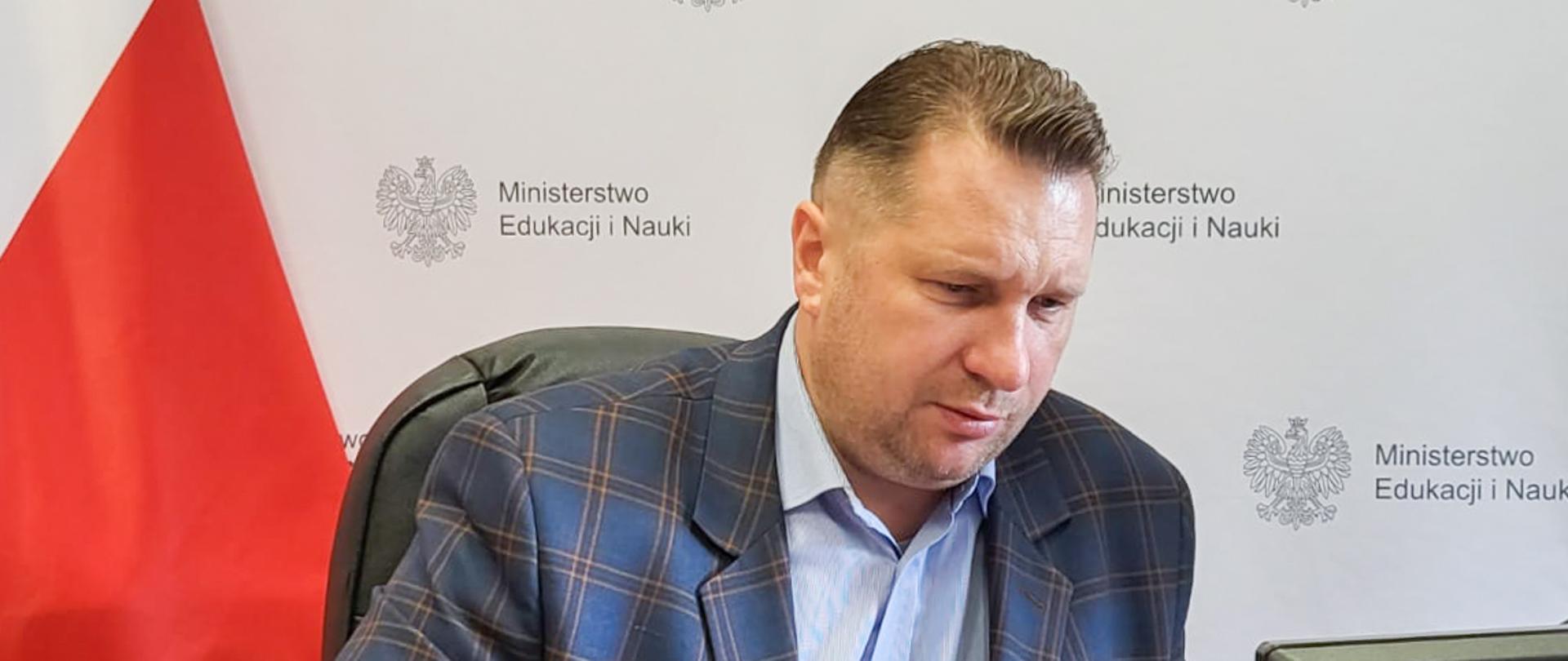 Minister Czarnek siedzi przy biurku, za nim polska flaga, przed nim monitor.
