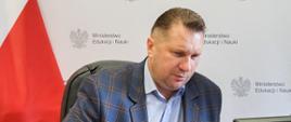 Minister Czarnek siedzi przy biurku, za nim polska flaga, przed nim monitor.