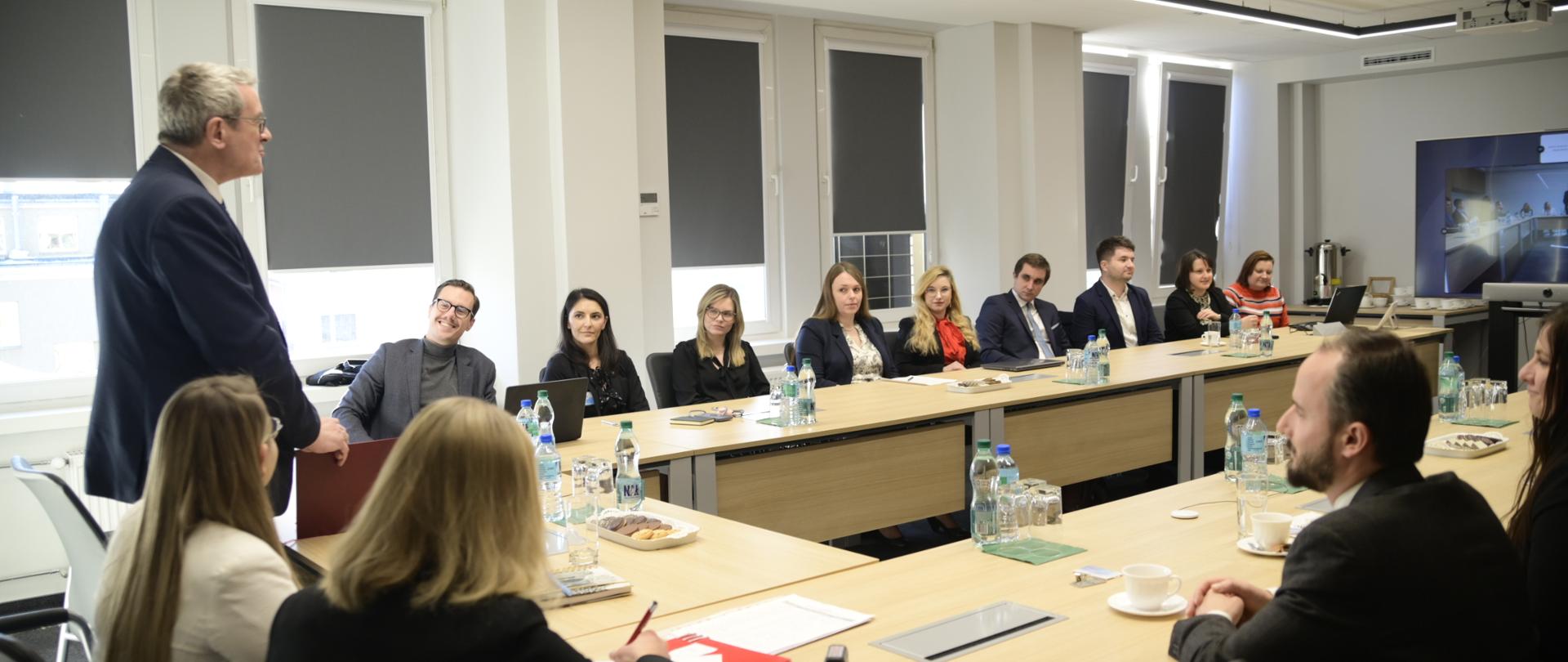 Na zdjęciu kilkanaście osób siedzących za stołem, jeden mężczyzna w eleganckim ubraniu stoi, w tle okna oraz ekran. 