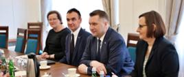 Przy drewnianym stole siedzi minister Czarnek, obok niego mężczyzna w garniturze i dwie ubrane na czarno kobiety, za nimi biała ściana i dwa okna.