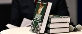 Na stoliku stoi książka oparta o leżące trzy książki. Obok leżą trzy mikrofony.