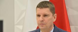 Za brązowym stołem siedzi wiceminister Piontkowski, za nim na żółtej ścianie wisi polska flaga.