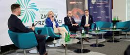 Wiceminister Machałek w niebieskiej marynarce siedzi na niebieskim fotelu i mówi do mikrofonu, na fotelach po obu stronach siedzi trzech mężczyzn w garniturach, za nimi ścianka z napisem Zawody przyszłości.