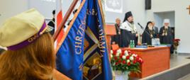 Widok na salę, na pierwszym planie poczet sztandarowy - dziewczyna w żółtej czapce ze sztandarem, dalej stół przy którym stoi mężczyzna w ceremonialnym czarnym ubraniu i mówi do mikrofonu.