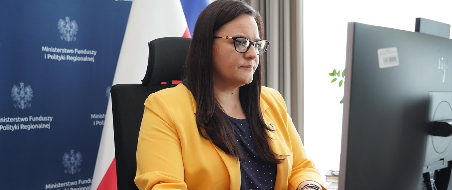 Wiceminister funduszy i polityki regionalnej Małgorzata Jarosińska-Jedynak podczas łączenia on-line na Konferencji Zero Project 2023, wiceminister siedzi przed komputerem, patrzy na ekran, za jej plecami flagi Polski i UE oraz baner MFiPR