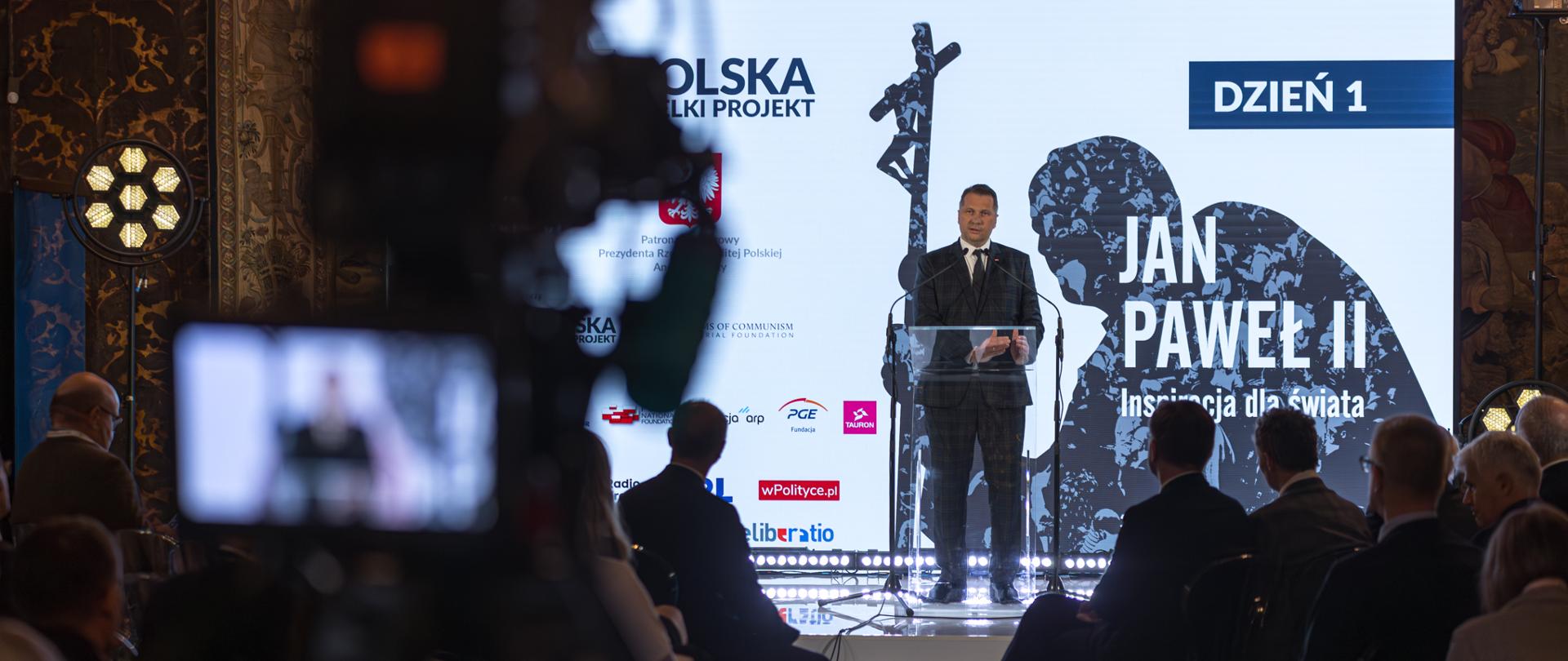 W zaciemnionej sali pod ekranem z napisem Dzień 1 - Jan Paweł II stoi minister Czarnek i mówi do mikrofonu na stojaku.