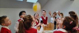 Dookoła stołu stoi grupa dzieci w białych koszulach i czerwonych kamizelkach, patrzą na unoszący się nad stołem żółty balonik.