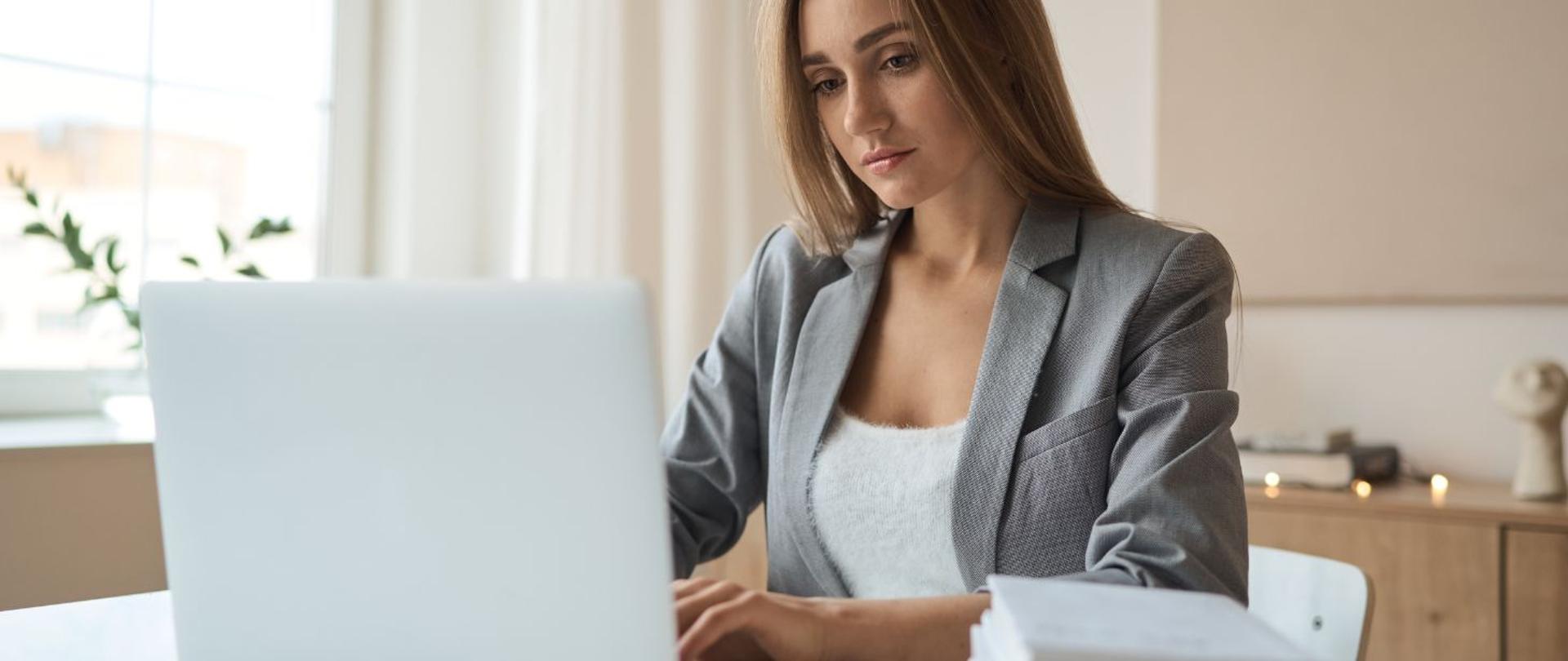 Informacja o pomocy skierowanej do małych i średnich przedsiębiorstw. Na zdjęciu elegancko ubrana kobieta pisze na laptopie i patrzy w ekran laptopa. W tle widok szafek biurowych i parapetu z kwiatkiem.