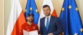 Minister Kamil Bortniczuk pozuje do zdjęcia z żużlowcem Bartoszem Zmarzlikiem. Minister ubrany jest w granatowy garnitur w kratkę, a żużlowiec w czerwoną bluzę reprezentacyjną z orzełkiem na piersi. Sportowiec trzyma w rękach list gratulacyjny otrzymany od ministra.