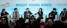 Na tle jasnoniebieskiej ściany z napisem Bright Young Minds w białych fotelach siedzi kilka osób, ubrana na ciemno kobieta mówi do mikrofonu, przed nimi widać sale na której siedzi wiele osób i słucha.