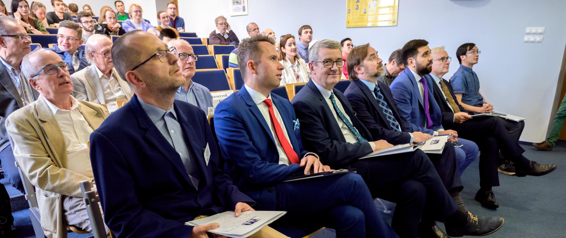 Zdjęcie pod kątem z boku, na ustawionych w rzędy krzesłach siedzi dużo ludzi, w pierwszym rzędzie pośrodku wiceminister Murdzek.
