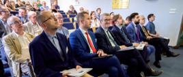 Zdjęcie pod kątem z boku, na ustawionych w rzędy krzesłach siedzi dużo ludzi, w pierwszym rzędzie pośrodku wiceminister Murdzek.