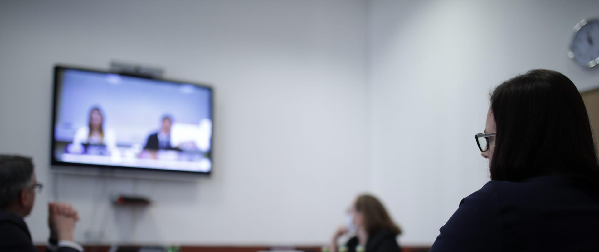Sylwetka minister siedzącej przy stole konferencyjnej, w tle telewizor i dwóch pracowników resortu. 